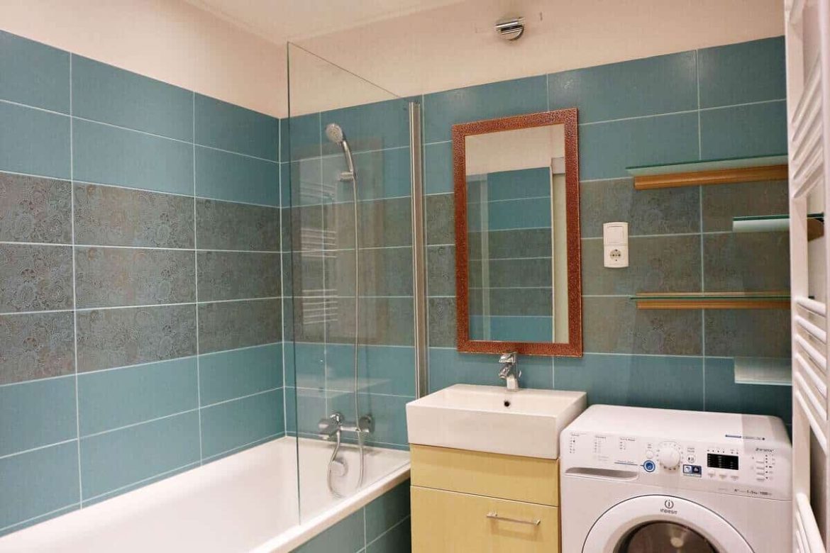 Une salle de bain moderne avec du carrelage bleu canard et gris texturé, comprenant une baignoire avec douche, un meuble sous-vasque jaune clair, un miroir et une machine à laver.