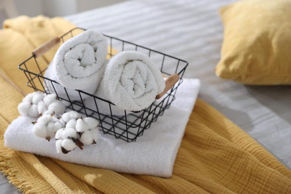 Panière métallique noire avec deux serviettes blanches roulées et branches de coton en fleur, reposant sur un plaid en gaze de coton jaune sur un lit, illustrant des articles de qualité issus d’un Déstockage Linge de Maison.