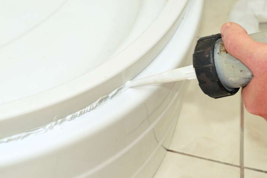 Une main applique un cordon de mastic silicone blanc à l'aide d'un pistolet extrudeur pour assurer l'étanchéité entre la paroi d'une baignoire et le carrelage mural blanc.